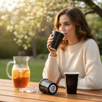 Woman drinking from a black CooCoo Cup outdoors with a pitcher of iced tea on a wooden table and a cup showing the bottom sticker featuring a "Brains, Beauty, and Booty" checklist-style design.