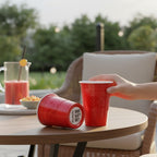 Red CooCoo Cups® on a table outdoors. A hand reaching out to one cup and another cup laying on its side showing a bottom sticker featuring a "Brains, Beauty, and Booty" checklist-style design.