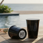 Two black CooCoo Cups® pool side on the beach. One cup displays the logo while the other lies on its side revealing the bottom sticker featuring "Beaches Booze Besties" text.
