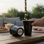 Two black CooCoo Cups® sit on a wooden outdoor table. A hand reaches for one cup, while the other lies on its side, revealing a bottom sticker featuring a classic 'BEER' emblem graphic.
