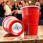 Red CooCoo Cups® with a classic "BEER" emblem bottom sticker on a wooden surface with people partying in the background.