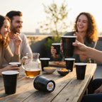 People enjoying a casual outdoor gathering with drinks in CooCoo Cups® on a wooden table. One cup shows the bottom sticker featuring two beer mugs clinking and “Cheers” text.