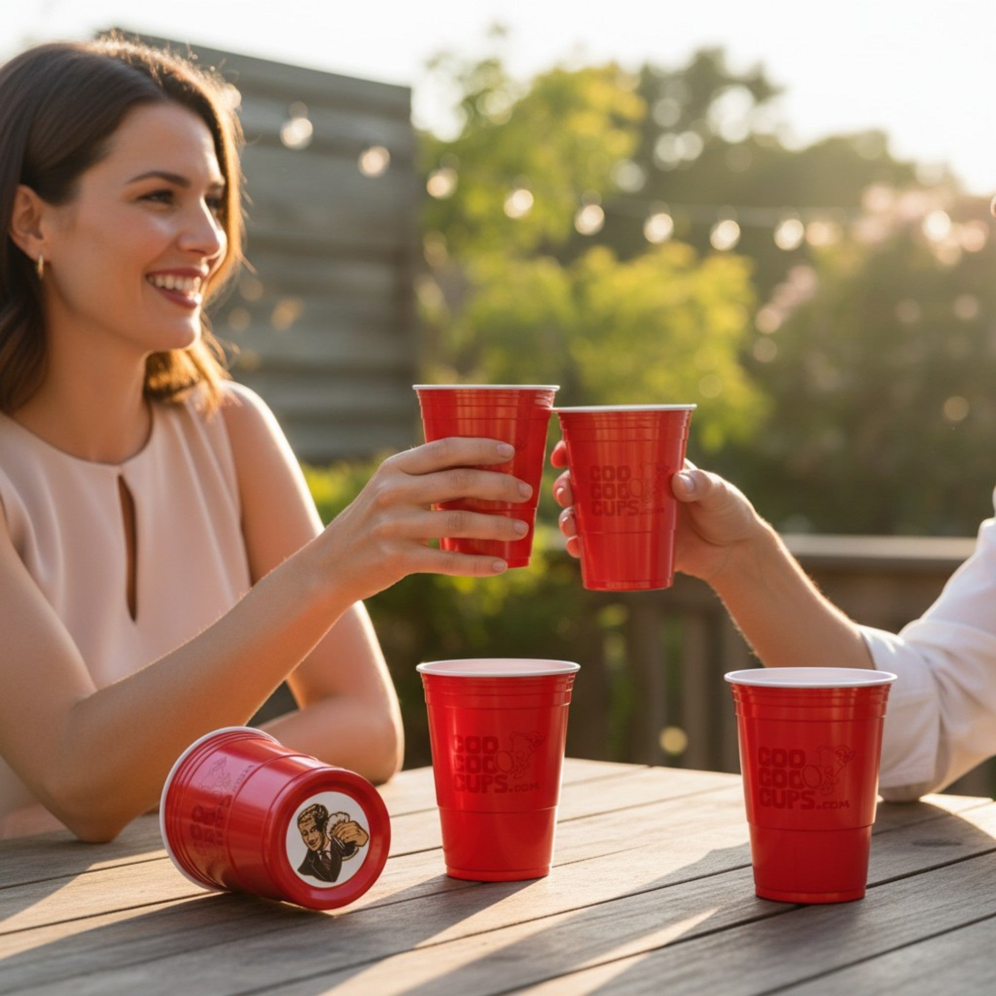 Two people toast with CooCoo Cups® on an outdoor patio. On the table, one cup lies on its side, revealing a bottom sticker graphic of a charismatic man holding a beer and raising a toast.