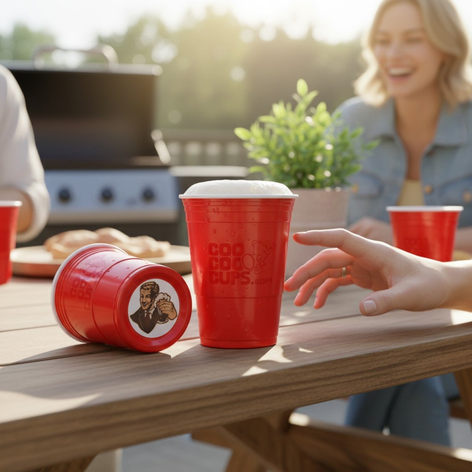 A fun outdoor barbecue scene features CooCoo Cups® on a picnic table with people enjoying themselves. A hand reaches for a cup of beer, while another cup lies on its side, revealing a bottom sticker graphic of a charismatic man holding a beer and raising a toast.