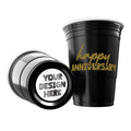 Two black CooCoo Cups® are shown. One displays the gold text “Happy Anniversary” while the other lies on its side, revealing a customizable bottom sticker labeled “Your Design Here.”