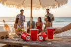 People enjoying a beach gathering with red CooCoo Cups® and snacks under an umbrella. Two cups laying down showing bottom stickers featuring "Beaches Booze Besties" and "Salty ASA Beach" graphics.