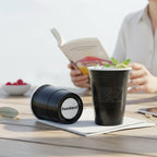 Two black CooCoo Cups® are shown on an outdoor table with a woman reading a book in the background. One displays the logo with a drink in it while the other lies on its side, revealing a bottom sticker labeled “#weekend.”