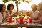 People enjoying dinner outside with CooCoo Cups®. Cups on the table lie on their sides, revealing bottom stickers featuring astronaut illustrations with peace-sign and dabbing poses.